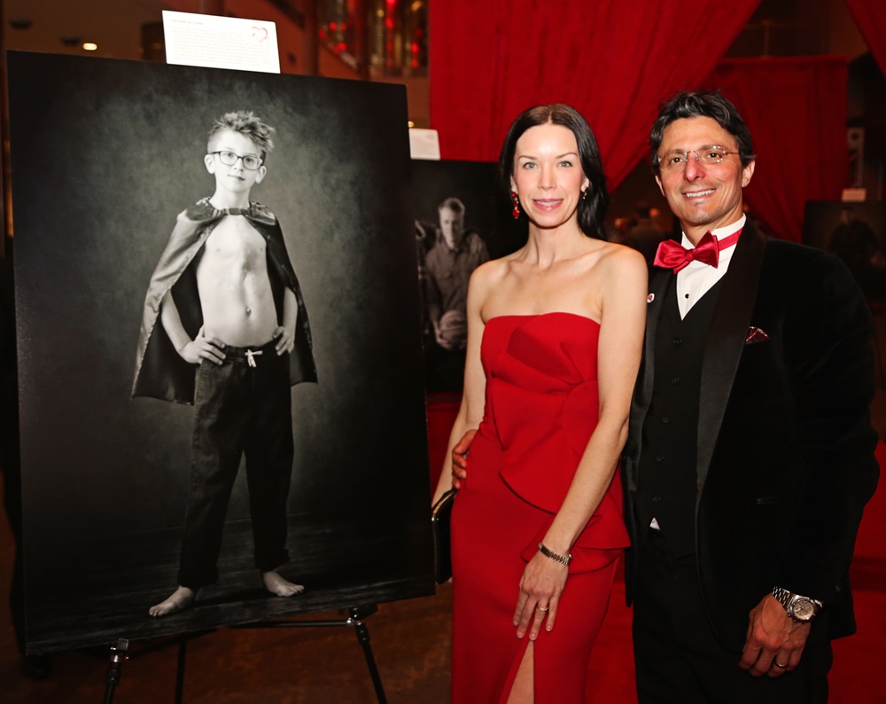 Couple in formal wear standing next to a black and white poster of a little boy with an open heart surgery scar.